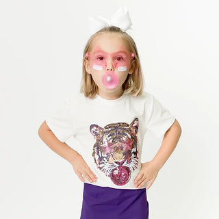 A young girl stands against a white studio background wearing a white graphic t-shirt with a colorful tiger face print, a purple mini skirt, and white sneakers. She has a white bow in her hair and is blowing a pink bubblegum bubble.