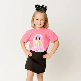 A young girl with blonde hair in a black bow stands against a white studio background, wearing a pink short-sleeve T-shirt with a large iridescent ghost graphic on the front, a black skirt, and white sneakers.