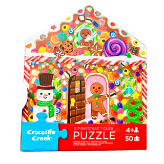 A hand holds a Crocodile Creek gingerbread house jigsaw puzzle box in a store setting. The packaging features a colorful Christmas gingerbread theme with a snowman, gingerbread people, candy canes, cookies, and a large red bottom section with the brand name and product details.