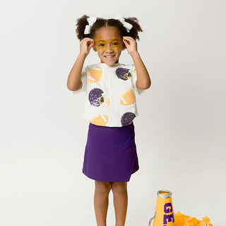 A young girl with pigtails stands on a white studio background wearing a white graphic T-shirt with orange and purple football patterns, a purple skirt, and white sneakers. She is holding a yellow megaphone with purple lettering that reads 'GEAUX' next to purple and orange pom-poms.